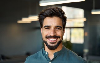 A man with dark hair and a beard wearing a dark green button-up and smiling