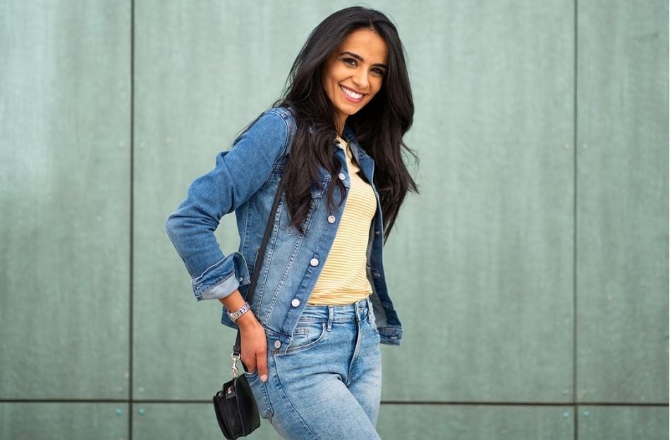 A woman with long, dark hair wearing a denim jacket and jeans smiling